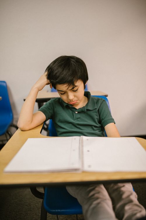 A young boy sitting alone at a desk, staring down with sadness
